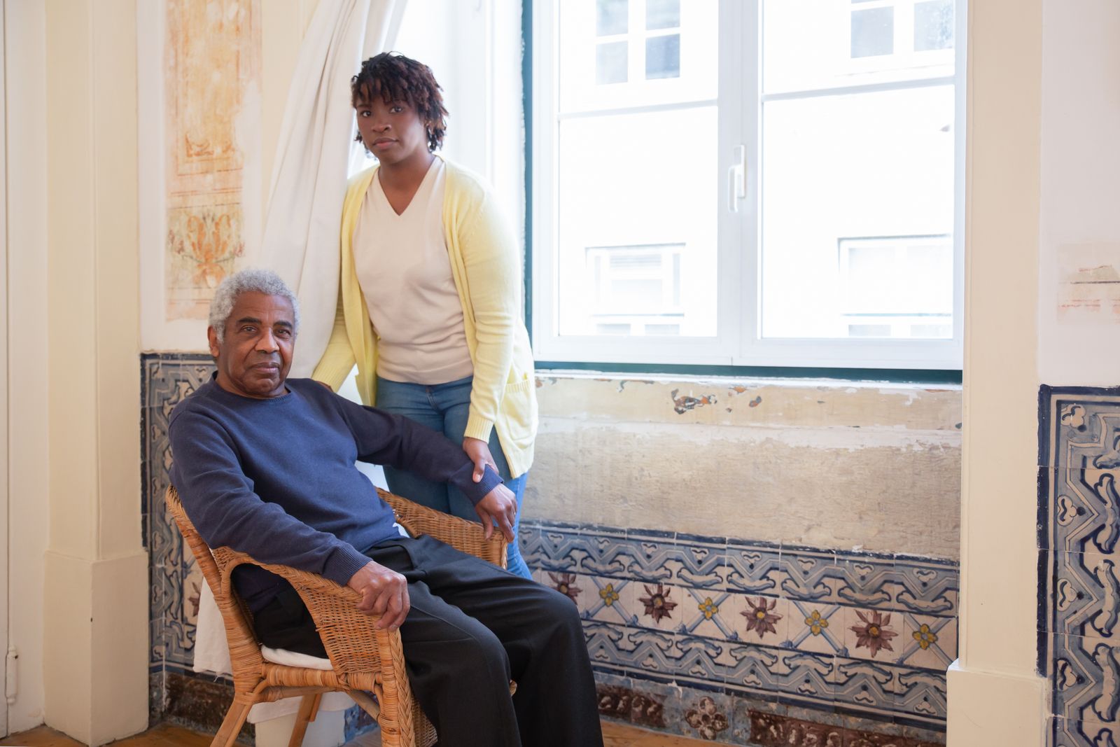 An older man seated by a window with a companion nearby